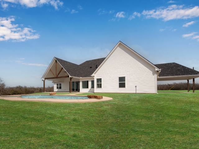 The Garrett Project&#039;s backyard features lush lawn, pool, and view of the white exterior of the house.