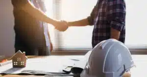 Two people shaking hands in front of a table with a hard hat and documents on top, choosing the right custom home builder.