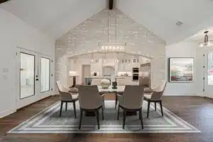 Living room with tall white brick walls, beige dining chairs on a white rug, pendant lights, and a white kitchen in the background.