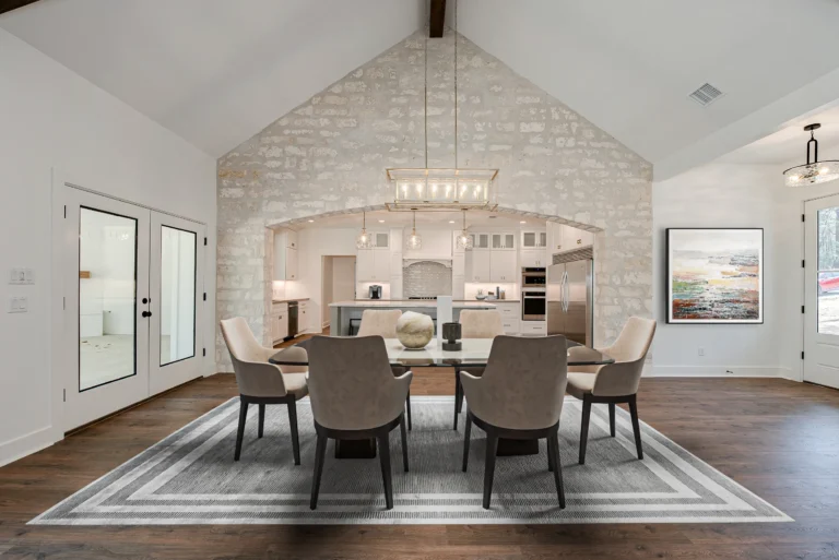 Living room with tall white brick walls, beige dining chairs on a white rug, pendant lights, and a white kitchen in the background.