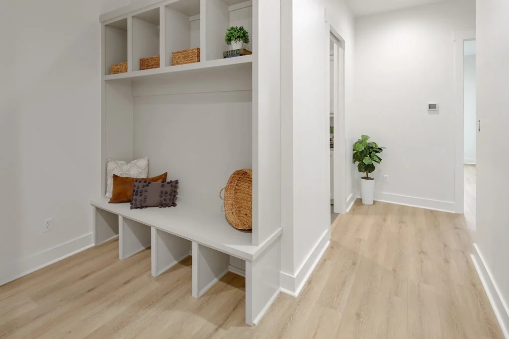 Current image: Guest-friendly home entryway with white cubby seating, decorative pillows, light wood flooring, and a corner plant